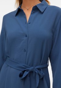 Robe chemise bleue à manches longues, avec boutons sur le devant, col pointu et ceinture en tissu nouée en nœud à la taille.
