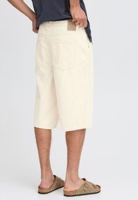 Pantalones cortos de algodón beige con un ajuste relajado, hasta la rodilla, que cuentan con bolsillos traseros y un parche texturizado de color marrón en la cinturilla. Combinados con sandalias marrones.