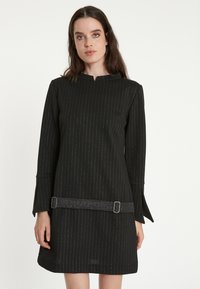 Schwarzes Kleid mit vertikalen silbernen Streifen, langen Ärmeln und einem subtilen Schlitz an den Bündchen. Verfügt über einen dekorativen, glitzernden Gürtel in der Taille.