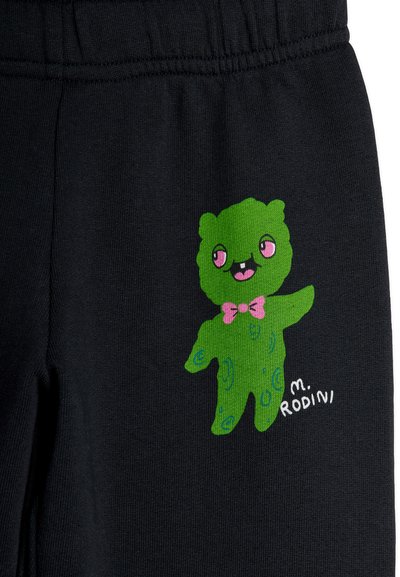 Πράσινος καρτούν αρκούδος με ροζ μάτια, ροζ παπιγιόν και μπλε βούλες σε μαύρο υφασμάτινο παντελόνι, με το κείμενο «M. Rodini» κοντά.