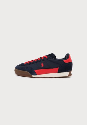 Baskets bleu marine avec des accents rouges, des lacets rouges, une semelle marron et un logo polo rouge sur le côté, présentées sur fond blanc uni.