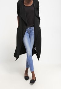 Abrigo negro largo con frente drapeado, llevado sobre una blusa negra y jeans azul claro. Zapatos planos grises completan el atuendo. Textura de tela suave.