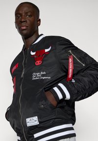 Fekete bomber dzseki piros Chicago Bulls logóval, fehér és fekete csíkozott bordás mandzsettákkal, hímzéssel és cipzáras zsebbel az ujjon.