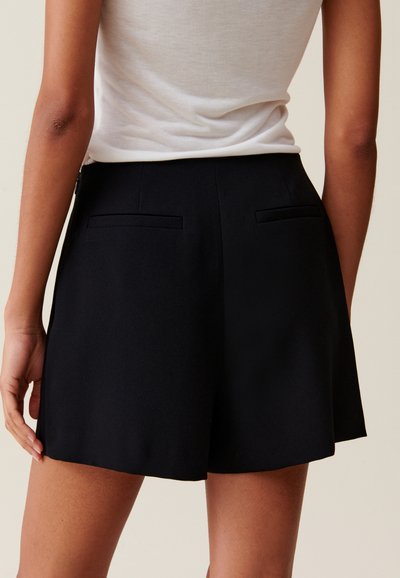 Shorts noirs cintrés avec une texture lisse, poches arrière plates et une silhouette épurée, associés à un haut ajusté de couleur claire.