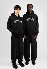Sudaderas con cremallera negra y texto gráfico blanco, combinadas con pantalones de mezclilla negra de corte holgado y zapatos negros. Textura suave y ajuste relajado.