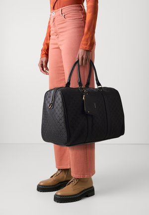 Sortmønstret weekendtaske med dobbelt håndtag, guldfarvede detaljer og en aftagelig mærke. Sammen med lyserøde jeans og tan farvede snørebåndsstøvler.