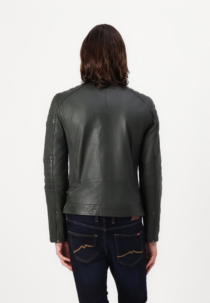 Homme aux cheveux mi-longs portant une veste en cuir gris foncé et un jean sombre, vu de dos contre un fond uni.