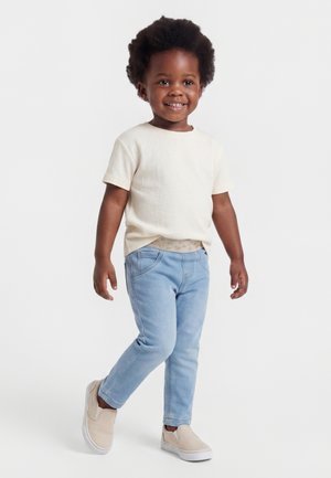 Tout-petit souriant portant un t-shirt crème à manches courtes, un jean bleu clair et des chaussures beiges à enfiler, debout sur un fond blanc.