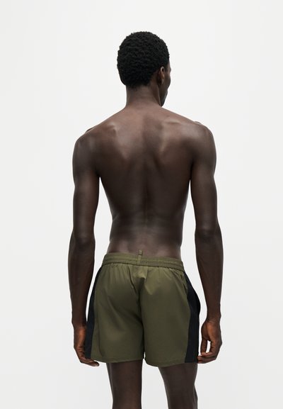 Un homme torse nu à la peau foncée portant un short vert olive et noir, debout de dos sur un fond clair uni.