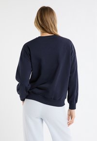 Felpa blu navy con maniche lunghe, polsini e orlo a costine, texture morbida. Indossata con pantaloni a quadri chiari. Vista posteriore.