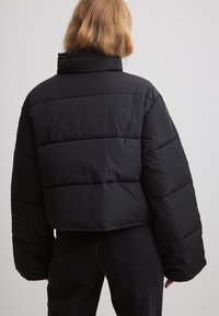 Melna apgriezta pūkota jaka ar platām piedurknēm, kurai ir kvadrātveida šuves. Gluda auduma un augsts apkakls, apvienota ar tumšām džinsu biksēm.