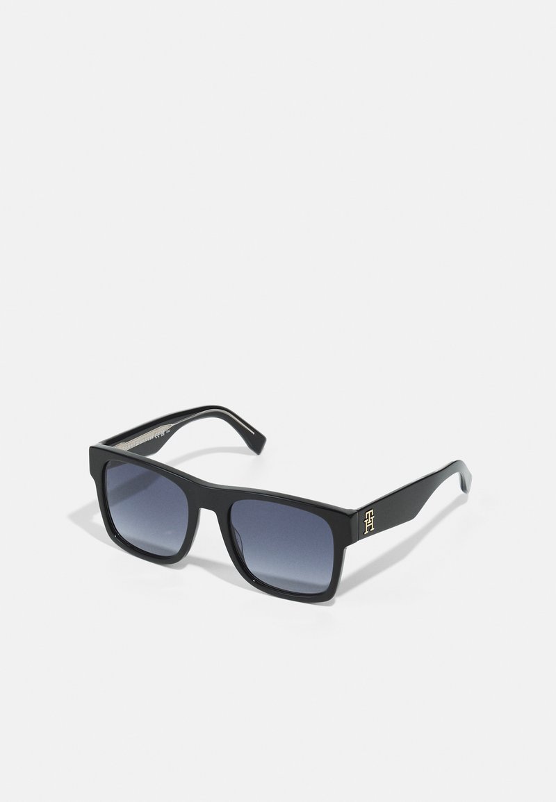 Lunettes de soleil noires rectangulaires avec des verres dégradés. Elles présentent un accent doré sur la branche et une monture en plastique lisse.