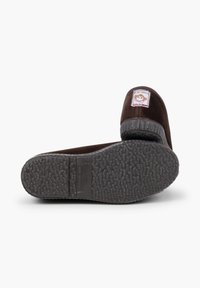 Pantofole in velluto marrone con suola in gomma testurizzata. Presentano una toppa circolare con logo sulla parte superiore, che indica l'origine "Made in Spain."
