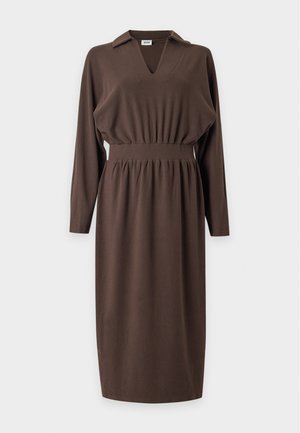 Femme debout portant une robe midi marron à manches longues avec un col et des sandales noires à talons hauts sur un fond blanc uni.