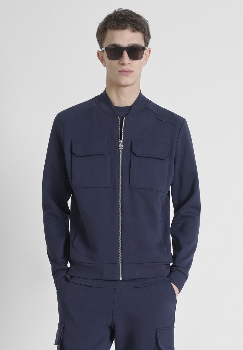 Chaqueta con cremallera en color navy, hecha de una tela suave, que presenta dos bolsillos en el pecho y un cuello acanalado. Combinada con pantalones a juego en el mismo color.
