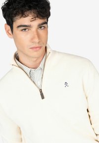 Suéter cremoso beige con cremallera y un patrón texturizado, con un logotipo azul marino. Llevado sobre una camisa de cuadro con cuello y cierre de botones.