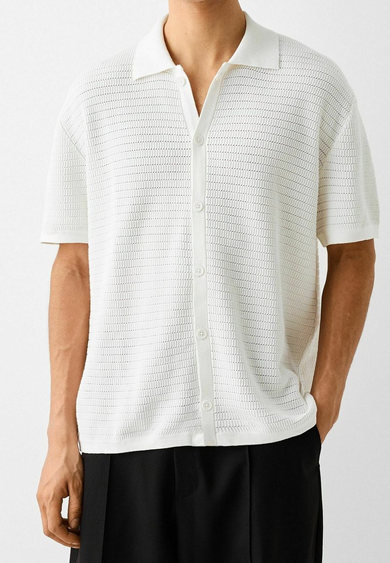 Homme portant une chemise en maille blanche à manches courtes boutonnée avec col et un pantalon noir, debout avec une main dans la poche.