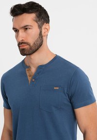 Modrá krátkosrstá tričko z měkkého materiálu s henley výstřihem a třemi knoflíky a kapsou na hrudi. Obsahuje značkový akcent.