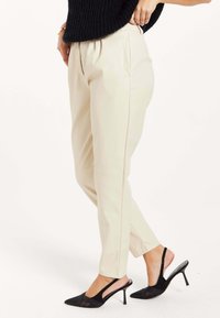 Beige faux leather trousers with a tapered fit, featuring front pleats, side pockets, and a smooth texture. Paired with black heeled shoes.