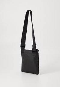 Sort crossbody taske med tekstureret stof, justerbar rem, lynlås lukning og logo detaljer på remmen. Kompakt og rektangulær form.