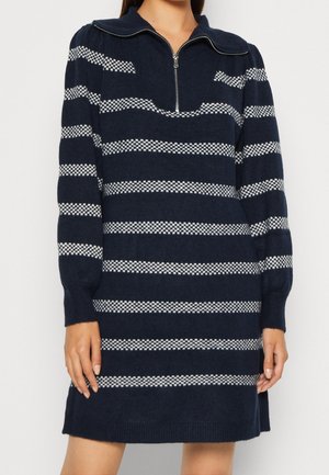 Femme portant une robe en maille bleu marine avec des rayures blanches horizontales à carreaux et un col à demi-fermeture éclair, debout devant un fond uni.