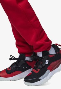 Zapatilla negra y roja con un upper texturizado, suela blanca y cordones negros, combinada con pantalones jogger rojos y un calcetín blanco.
