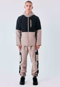 Chaqueta bicolor para hombre en negro y beige, combinada con pantalones a juego. Cuenta con bolsillos con cremallera y costuras reforzadas. Fabricada con tejido duradero.
