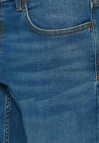 Jeans de mezclilla en azul medio, con un diseño clásico de cinco bolsillos, costuras visibles en naranja y una textura de tela suave.