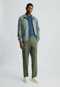 Die leichte Herrenjacke hat ein karriertes Muster in Grün und Blau, wird über einem blauen Poloshirt getragen und ist kombiniert mit olivgrünen Hosen und weißen Sneakern.
