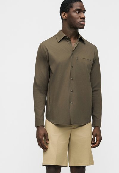 Homme debout portant une chemise boutonnée vert olive à manches longues et un short beige arrivant aux genoux, devant un fond uni.