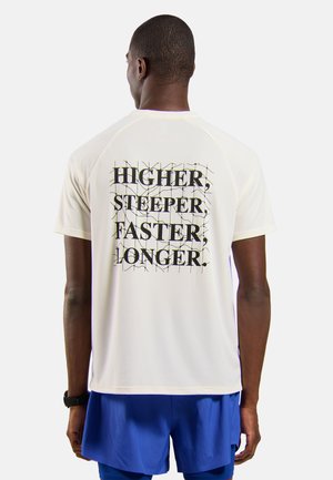 Man draagt een wit T-shirt met korte mouwen met zwarte tekst "HIGHER, STEEPER, FASTER, LONGER." op de rug en blauwe korte broek, staand met de rug naar het beeld.