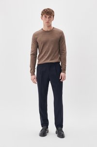 Barna kötött pulóver hosszú ujjakkal, formális sötétkék nadrággal párosítva. A modell egy sima fehér háttér előtt áll, fekete slip-on cipőt visel.