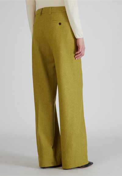 Vrouw in olijfgroene hoge taille, wijde broek met knoopzakken aan de achterkant en zwarte puntige schoenen, staand tegen een effen lichte achtergrond.