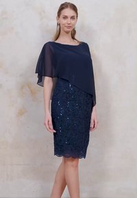 Mörkblå klänning med en lager av chiffonkappa, paljetterad textur, figurnära silhuett och vågig fåll. Elegant och formell design.