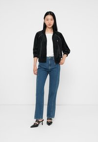 Chaqueta bomber de terciopelo negro con dos bolsillos laterales, llevada sobre una blusa blanca, combinada con vaqueros de cintura alta azules y tacones negros de punta.