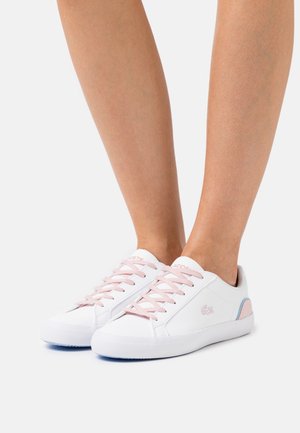 Pieds chaussés de baskets blanches à coupe basse avec des lacets roses et des accents bleus, posés sur un fond blanc.