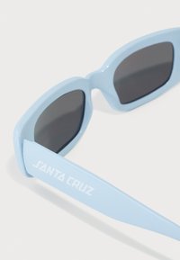 Gafas de sol azul cielo con lentes negros, diseño angular y patillas gruesas que presentan el texto blanco "SANTA CRUZ". Textura de plástico suave.