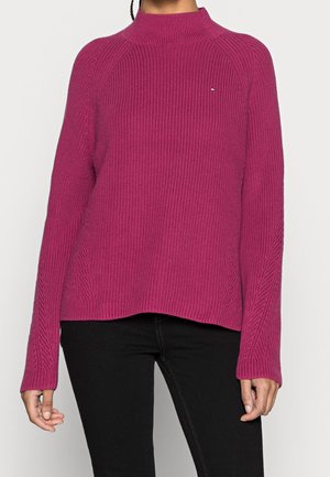 Femme portant un pull à col roulé rose côtelé à manches longues avec un petit logo sur la poitrine et un pantalon noir.