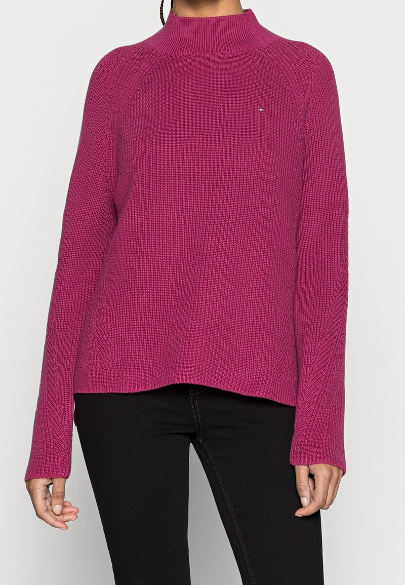 Femme portant un pull à col roulé rose côtelé à manches longues avec un petit logo sur la poitrine et un pantalon noir.