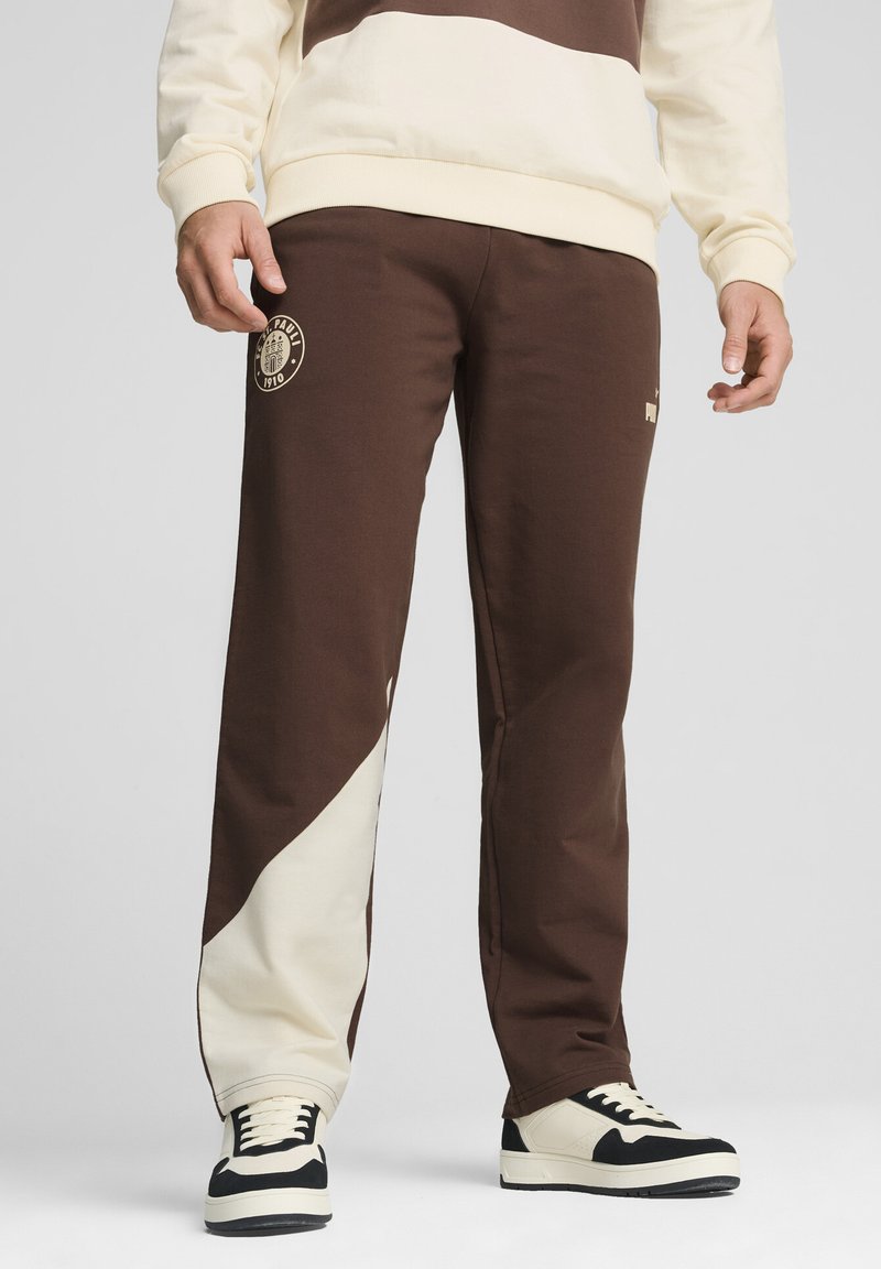 Braune und cremefarbene Sweatpants mit einem diagonalen cremefarbenen Einsatz. Verfügt über ein Logo-Patch und ein tapered Design, kombiniert mit schwarz-weißen Sneakers.