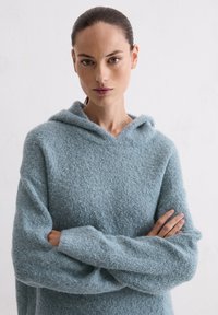 Jasnoniebieski, teksturowany sweter z kapturem wykonany z miękkiego materiału, z długimi rękawami i luźnym fasonem. Brak widocznych wzorów lub elementów metalowych.