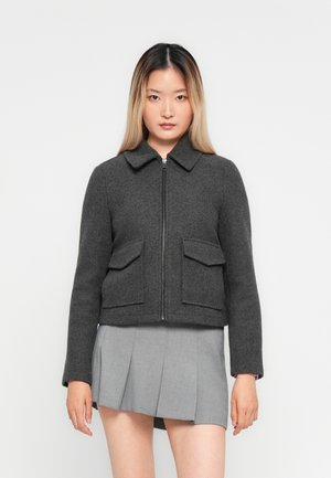 Žena s dugom ombre kosom koja nosi tamnosivu jaknu na patent s preklopnim džepovima i svijetlosivu plisiranu mini suknju na bijeloj pozadini.