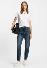 Witte T-shirt met korte mouwen en zwart "HUGO" logo, gecombineerd met hoge blauwe denim jeans. Model houdt een zwarte handtas vast en draagt chunky witte sneakers.