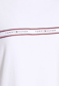 Bílá bavlněná tričko s horizontálním pruhovaným páskem v červené a modré barvě s nápisem „TOMMY HILFIGER“. Hladká textura, design s kulatým výstřihem.