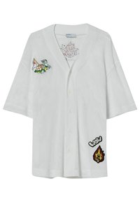 Valkoinen verkkomallinen neuletakki lyhyillä hihoilla, jossa vihreät kilpikonna- ja lippupaikat sekä mustat ja punaiset "WOW"- ja liekkikuvioinnit.