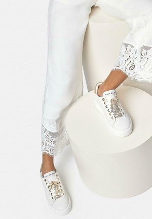 Persona con pantalones blancos con encaje en los bordes y zapatillas blancas con adornos de escorpiones dorados y plateados, descansando un pie sobre un taburete blanco redondo.