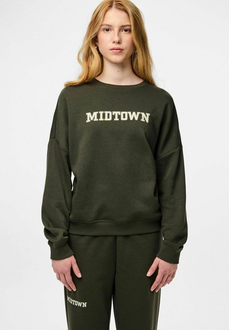 Jauna moteris su ilgais blondiniais plaukais, vilkinti tamsiai žalią džemperio ir kelnių komplektą, ant kurių šviesiomis raidėmis užrašyta „MIDTOWN“.