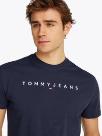 Marineblå bomulls-t-skjorte med "TOMMY JEANS" brodert i hvitt, med en liten rød og hvit logo. Klassisk rund hals og korte ermer.