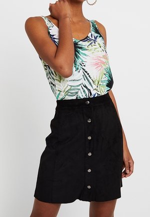 Femme portant un haut sans manches à imprimé feuilles tropicales et une jupe noire boutonnée devant, posant avec une main près du cou.
