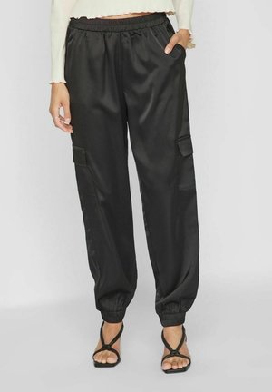 Femme portant un pantalon cargo en satin noir avec des poignets élastiques, associé à des talons noirs à lanières et un haut court crème à manches longues.